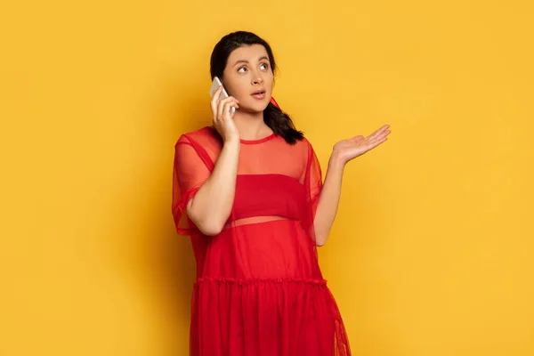 Mujer embarazada confundida en traje rojo que muestra gesto de encogimiento mientras habla en el teléfono inteligente en amarillo - foto de stock