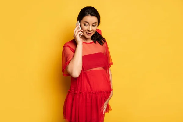 Jeune femme enceinte en tunique rouge toucher le ventre tout en parlant sur smartphone sur jaune — Photo de stock