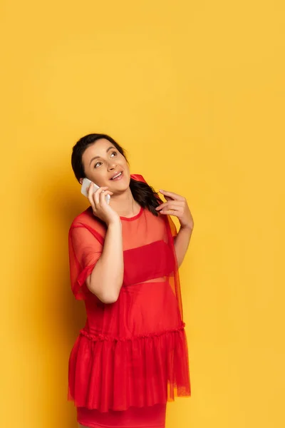 Mujer embarazada joven en túnica roja mirando hacia arriba mientras habla en el teléfono inteligente en amarillo - foto de stock