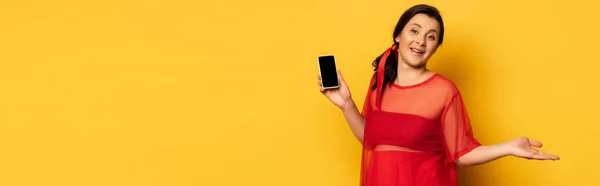 Вагітна жінка в червоному вбранні тримає смартфон з порожнім екраном на жовтому, заголовок веб-сайту — стокове фото