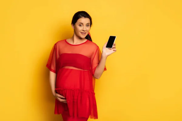 Брюнетка вагітна жінка в червоній туніці, що показує смартфон з порожнім екраном, торкаючись животика на жовтому — стокове фото