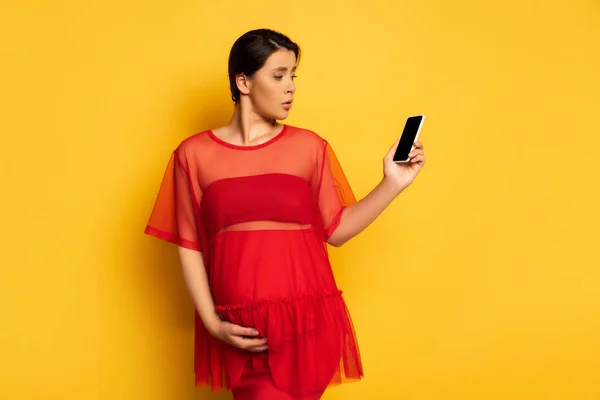 Bouleversé femme enceinte n tunique rouge tenant smartphone avec écran blanc tout en touchant le ventre sur jaune — Photo de stock