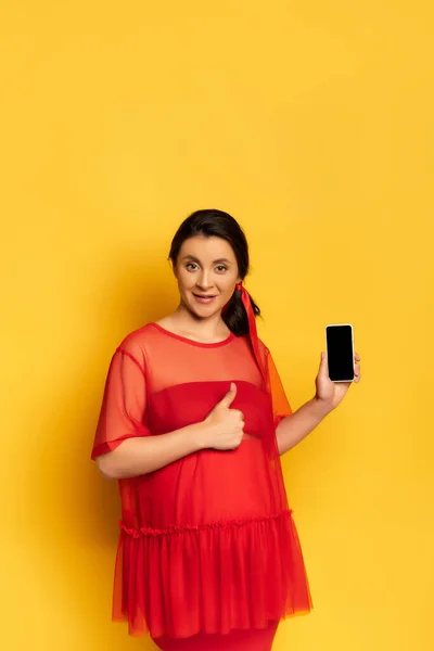 Pregnant woman in red outfit showing thumb up while holding smartphone with blank screen on yellow — Stock Photo