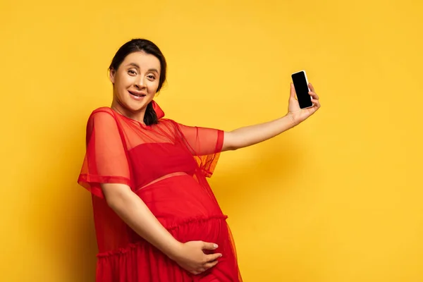 Mulher grávida morena em túnica vermelha levando selfie no smartphone enquanto olha para a câmera no amarelo — Fotografia de Stock