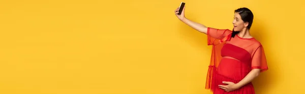 Horizontal image of pregnant woman in red tunic taking selfie on smartphone on yellow — Stock Photo