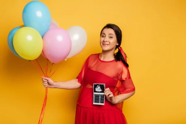 Pregnant woman in red outfit holding colorful festive balloons while showing ultrasound scan on yellow — Stock Photo