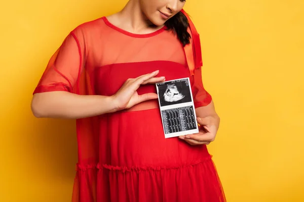 Teilaufnahme einer schwangeren Frau in rotem Outfit mit Ultraschalluntersuchung in Bauchnähe auf gelb — Stockfoto