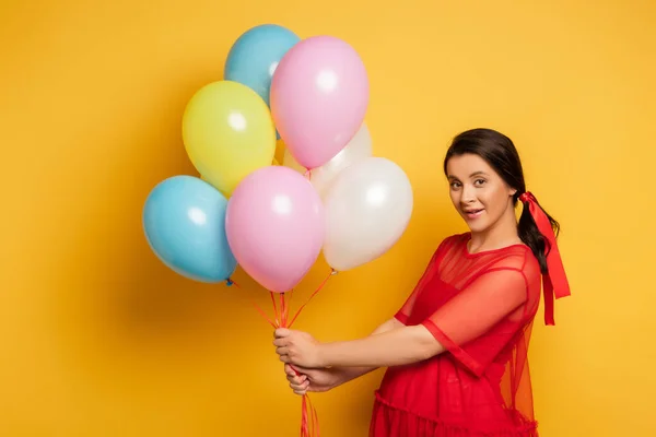 Brünette schwangere Frau im roten Outfit blickt in die Kamera, während sie bunte festliche Luftballons auf gelb hält — Stockfoto