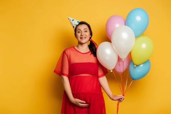 Young pregnant woman in party cap holding colorful festive balloons on yellow — Stock Photo