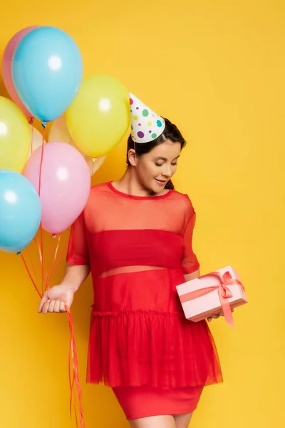Молодая беременная женщина в шапочке для вечеринок держит красочные праздничные воздушные шары и держит подарочную коробку на желтом — стоковое фото