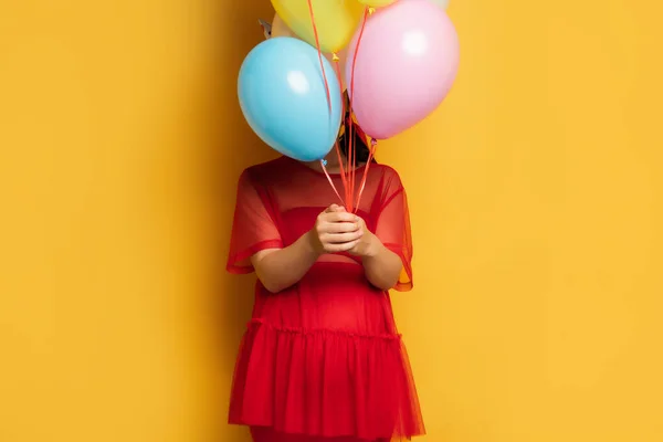 Giovane donna incinta in abito rosso viso oscuro con palloncini festivi colorati su giallo — Foto stock