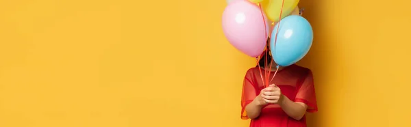 Горизонтальне зображення вагітної жінки в червоній туніці затемнює обличчя різнокольоровими святковими кульками на жовтому — стокове фото