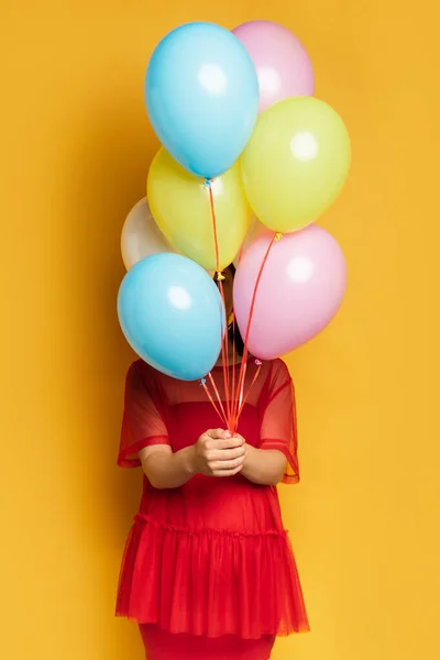 Schwangere in rotem Outfit verdeckt Gesicht mit bunten Luftballons auf gelb — Stockfoto