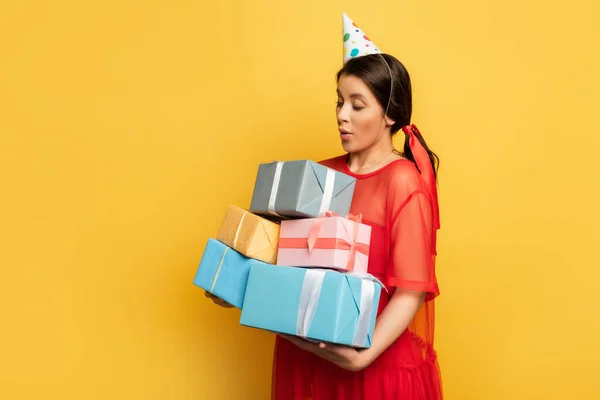 Удивленная беременная женщина в кепке для вечеринок держит стопку подарочных коробок на желтом — стоковое фото