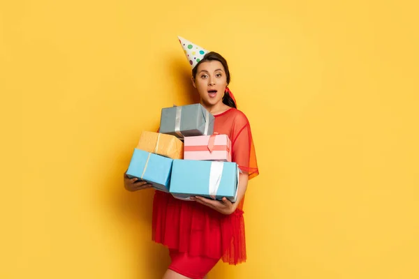 Shocked pregnant woman in red tunic holding stack of gifts while looking at camera on yellow — Stock Photo