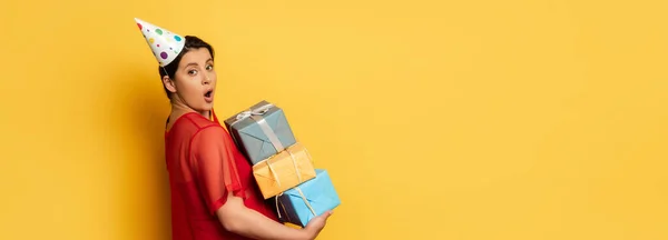 Horizontal image of surprised pregnant woman in party car holding stack of presents while looking at camera on yellow — Stock Photo