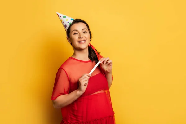 Pregnant woman in party cap looking at camera and holding pregnancy test on yellow — Stock Photo