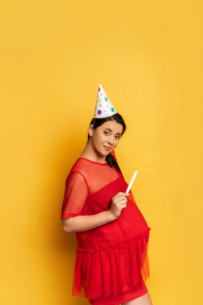 Mujer embarazada con gorra de fiesta que muestra la prueba de embarazo mientras mira a la cámara en amarillo - foto de stock