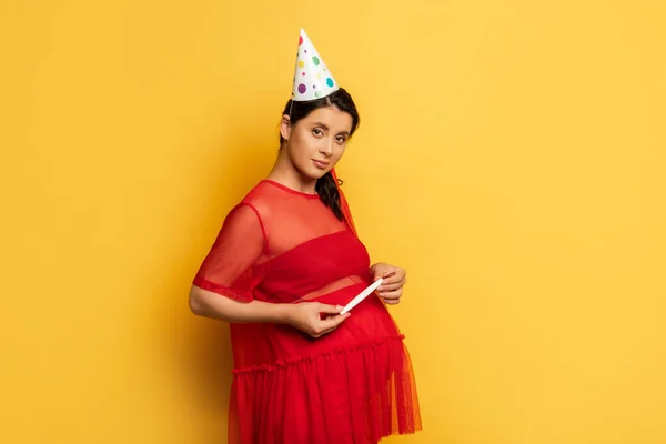 Mujer embarazada joven con gorra de fiesta y traje rojo que sostiene la prueba de embarazo en amarillo - foto de stock