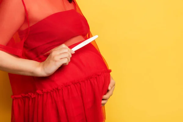 Vue recadrée de la femme enceinte en tenue rouge tenant test de grossesse tout en touchant le ventre sur jaune — Photo de stock