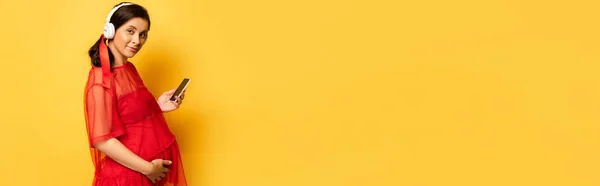 Conceito horizontal de mulher grávida em fones de ouvido sem fio tocando barriga e segurando smartphone no amarelo — Fotografia de Stock