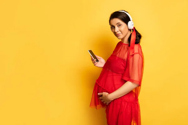 Mulher grávida em fones de ouvido sem fio segurando smartphone enquanto toca na barriga no amarelo — Fotografia de Stock
