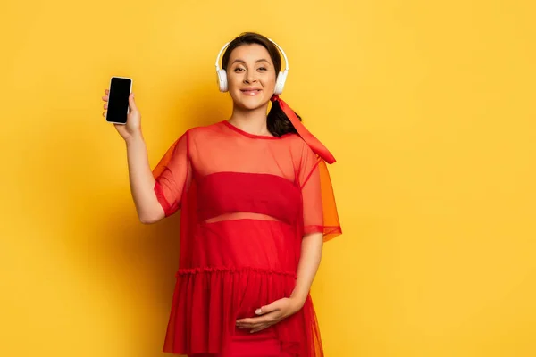 Pregnant woman in wireless headphones showing smartphone while touching tummy on yellow — Stock Photo