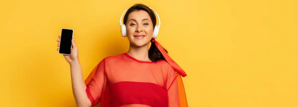 Image horizontale de la femme dans les écouteurs sans fil montrant smartphone avec écran blanc sur jaune — Photo de stock