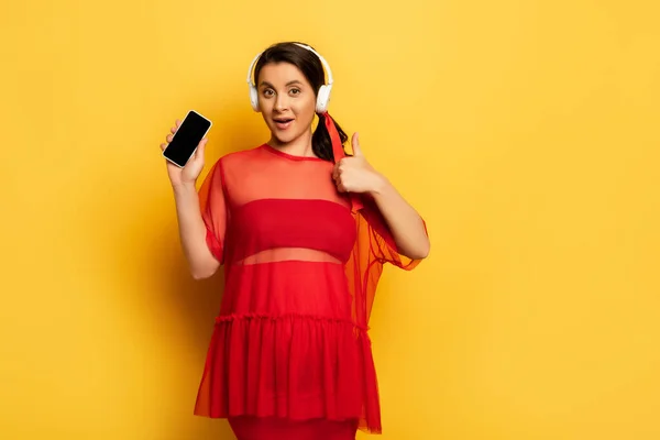 Mujer joven en túnica roja que muestra el teléfono inteligente con la pantalla en blanco y el gesto bien en amarillo - foto de stock