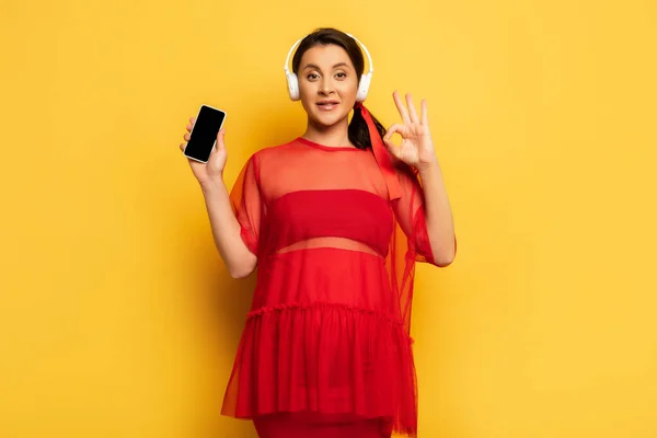 Donna incinta in cuffie wireless mostrando smartphone e gesto ok sul giallo — Foto stock