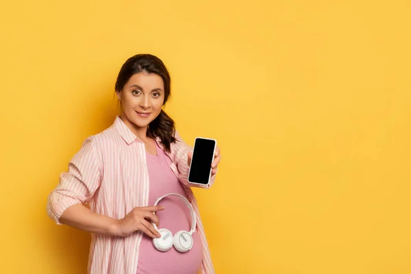 Frau zeigt Smartphone mit leerem Bildschirm und hält kabellosen Kopfhörer in Bauchnähe auf gelb — Stockfoto