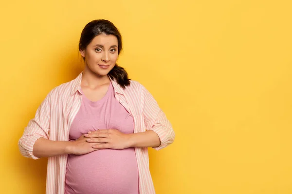 Молода вагітна жінка торкається живота, дивлячись на камеру на жовтому — стокове фото