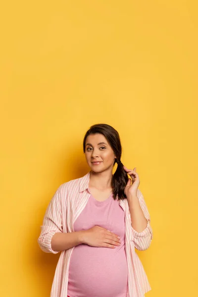 Donna incinta che tocca pancia e capelli mentre guarda la fotocamera su giallo con spazio copia — Foto stock
