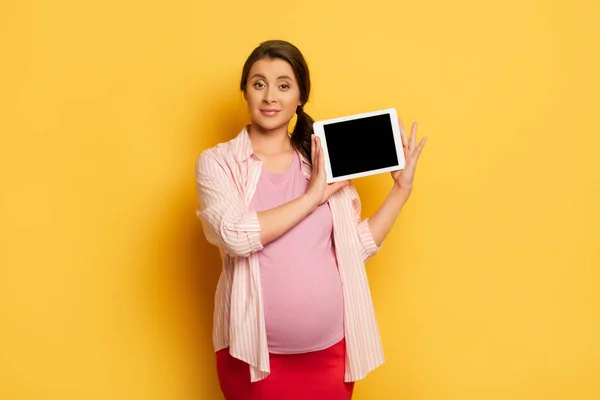 Беременная женщина смотрит в камеру, показывая цифровой планшет с чистым экраном на желтом — стоковое фото