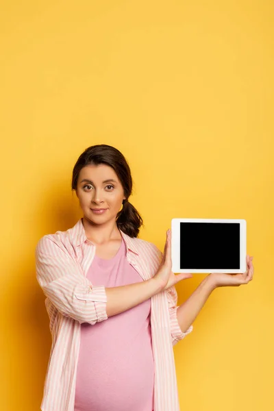 Pregnant woman showing digital tablet with blank screen and looking at camera on yellow — Stock Photo