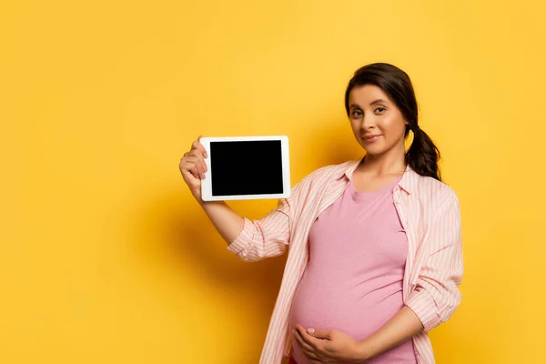 Pregnant woman touching belly while showing digital tablet with blank screen on yellow — Stock Photo