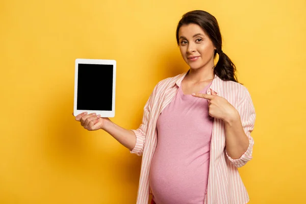 Вагітна жінка вказує на цифровий планшет з порожнім екраном на жовтому — стокове фото