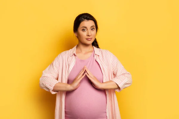Jeune femme enceinte montrant un geste de protection près du ventre tout en regardant la caméra sur jaune — Photo de stock