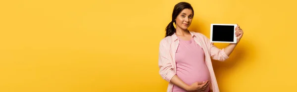 Horizontal image of young pregnant woman showing digital tablet with blank screen on yellow — Stock Photo
