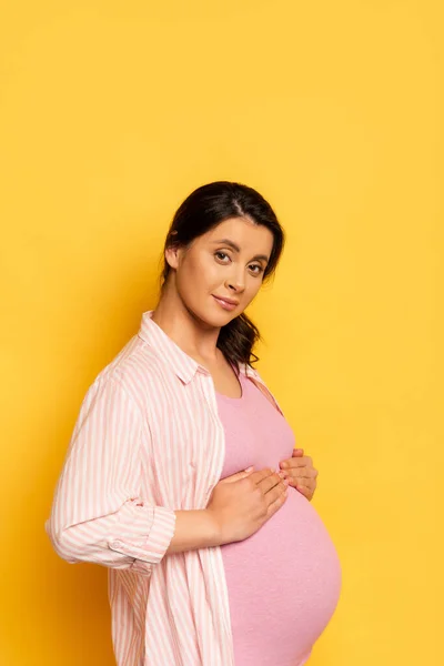 Young pregnant woman touching belly while looking at camera on yellow — Stock Photo
