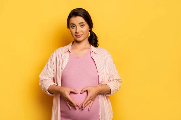 Brunette femme enceinte montrant le symbole du cœur près du ventre tout en regardant la caméra sur jaune — Photo de stock