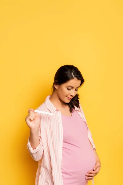 Embarazada mujer joven sosteniendo prueba de embarazo mientras toca la panza en amarillo - foto de stock