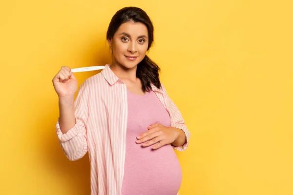 Беременная женщина трогает живот и показывает тест на беременность на желтом — стоковое фото