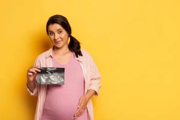 Pregnant woman showing ultrasound scan while touching belly on yellow — Stock Photo