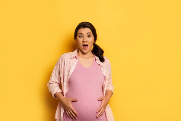Schockierte Schwangere blickt in Kamera, während sie Bauch auf gelb berührt — Stockfoto
