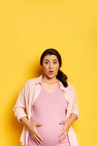 Sorprendida mujer embarazada tocando la barriga y mirando a la cámara con la boca abierta en amarillo - foto de stock