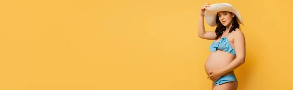 Horizontales Bild im Badeanzug, der Bauch und Strohhut berührt, während er auf gelb posiert — Stockfoto
