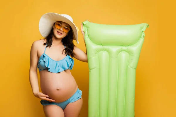 Pregnant woman touching belly while posing with inflatable mattress on yellow — Stock Photo