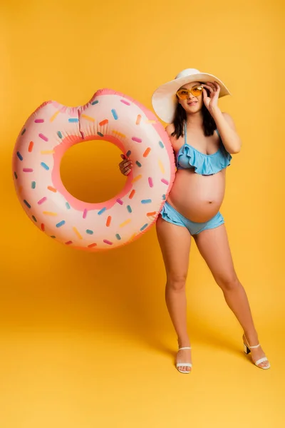 Young pregnant woman touching sunglasses while holding swim ring on yellow — Stock Photo