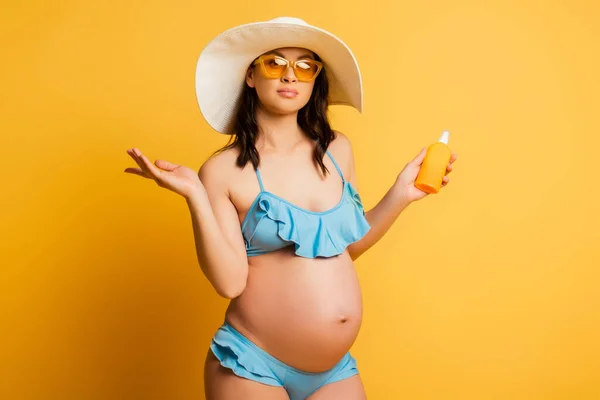 Беременная женщина в купальнике и соломенной шляпе держит солнцезащитный крем на желтом — стоковое фото